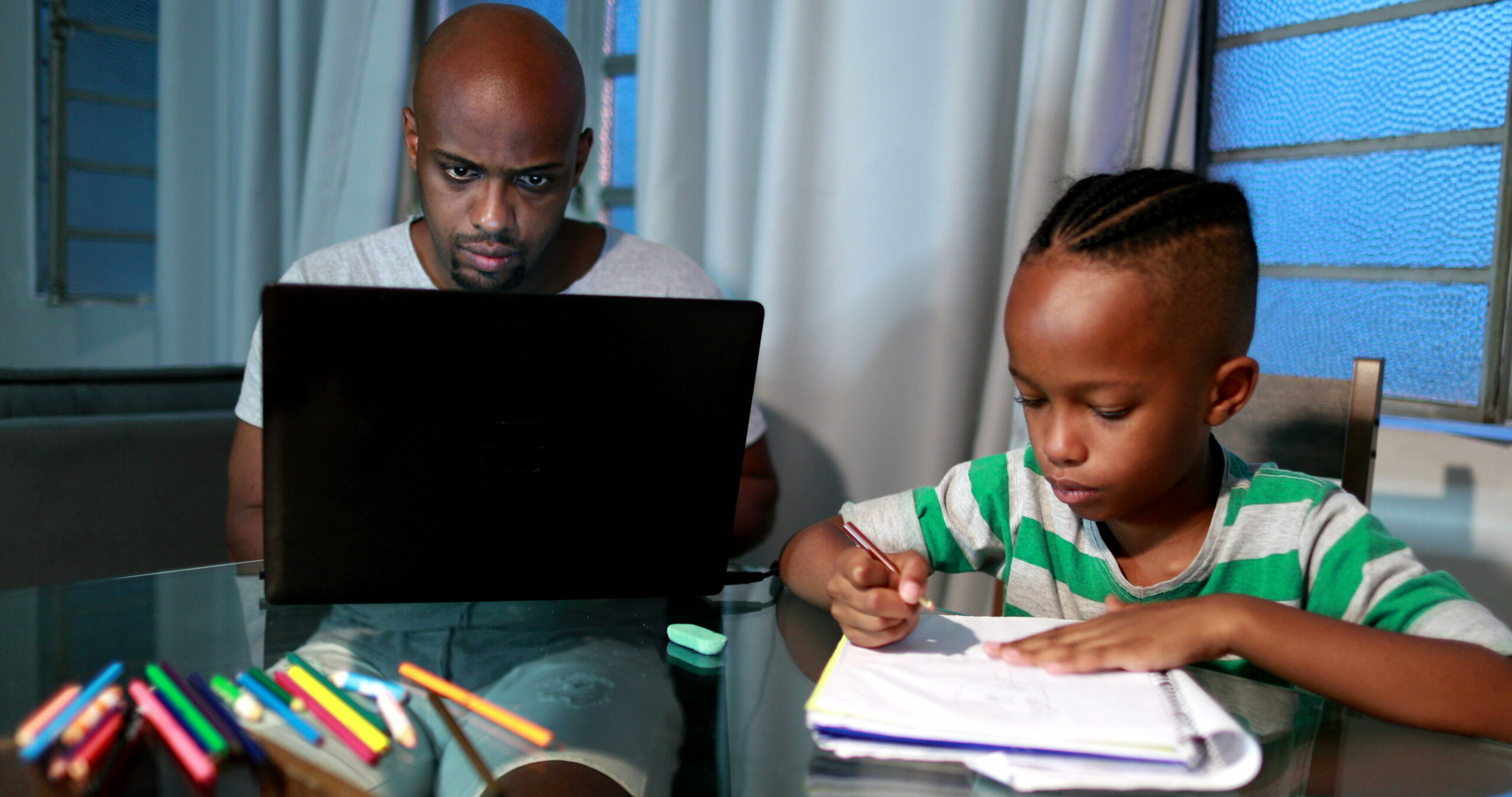 Father working on his laptop while his son draws on paper next to him